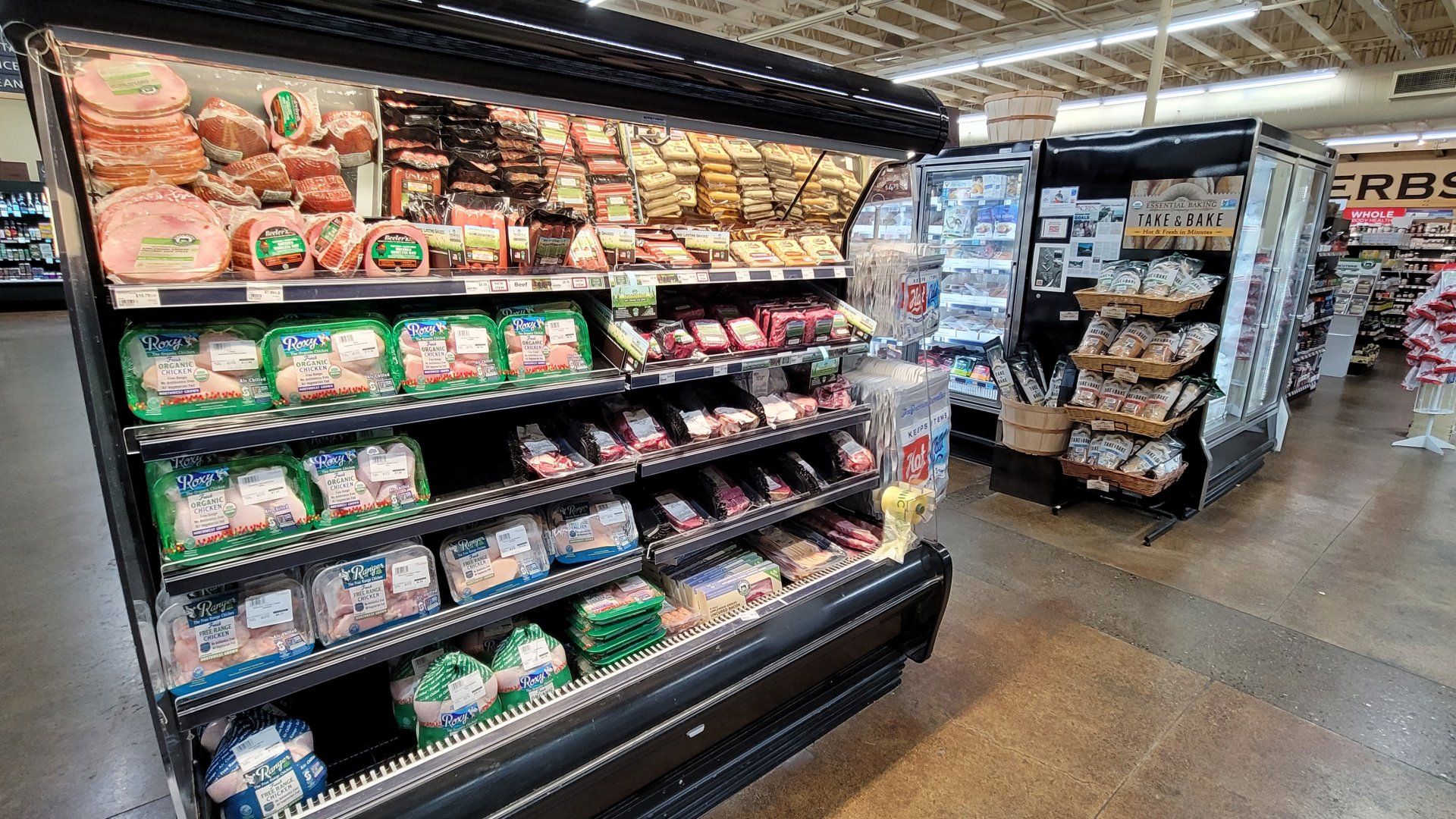 A refrigerated display case in a grocery store, filled with various packaged meats like ham and turkey. Another display case is nearby.