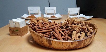 A wooden bowl filled with cinnamon sticks, cookies, and other cinnamon products on a wooden table. There are sample powders in white dishes.