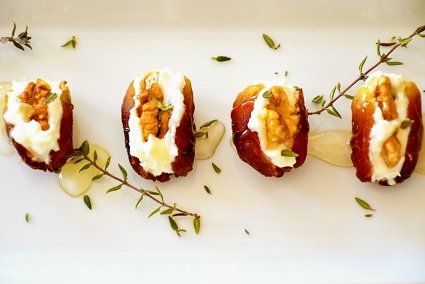 Four dates stuffed with creamy filling and walnuts, garnished with herbs and a drizzle of sauce on a white plate.
