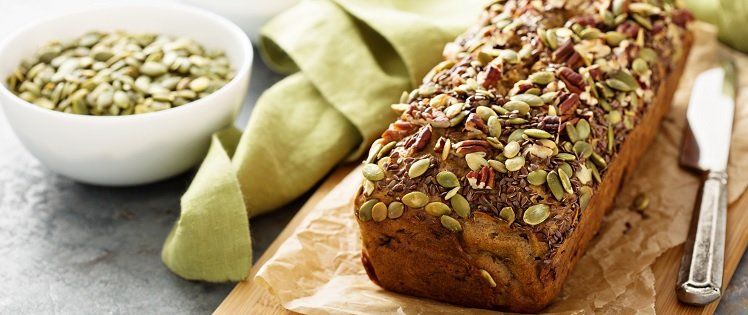 A loaf of seed-covered bread on a wooden board, next to a bowl of seeds and a butter knife. Green cloth in background.