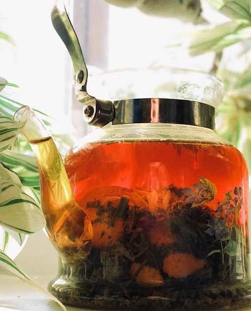 A glass teapot steeps a reddish-brown tea, with visible herbs, set near a window with leafy plants.
