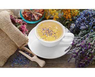 A cup of herbal tea with dried flowers, surrounded by various herbs and a wooden scoop on a wooden table.