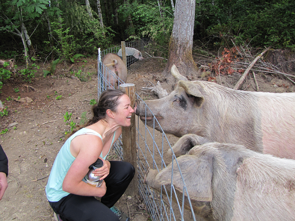 Woman smiles, touching snouts with pigs through a fence in a wooded area.
