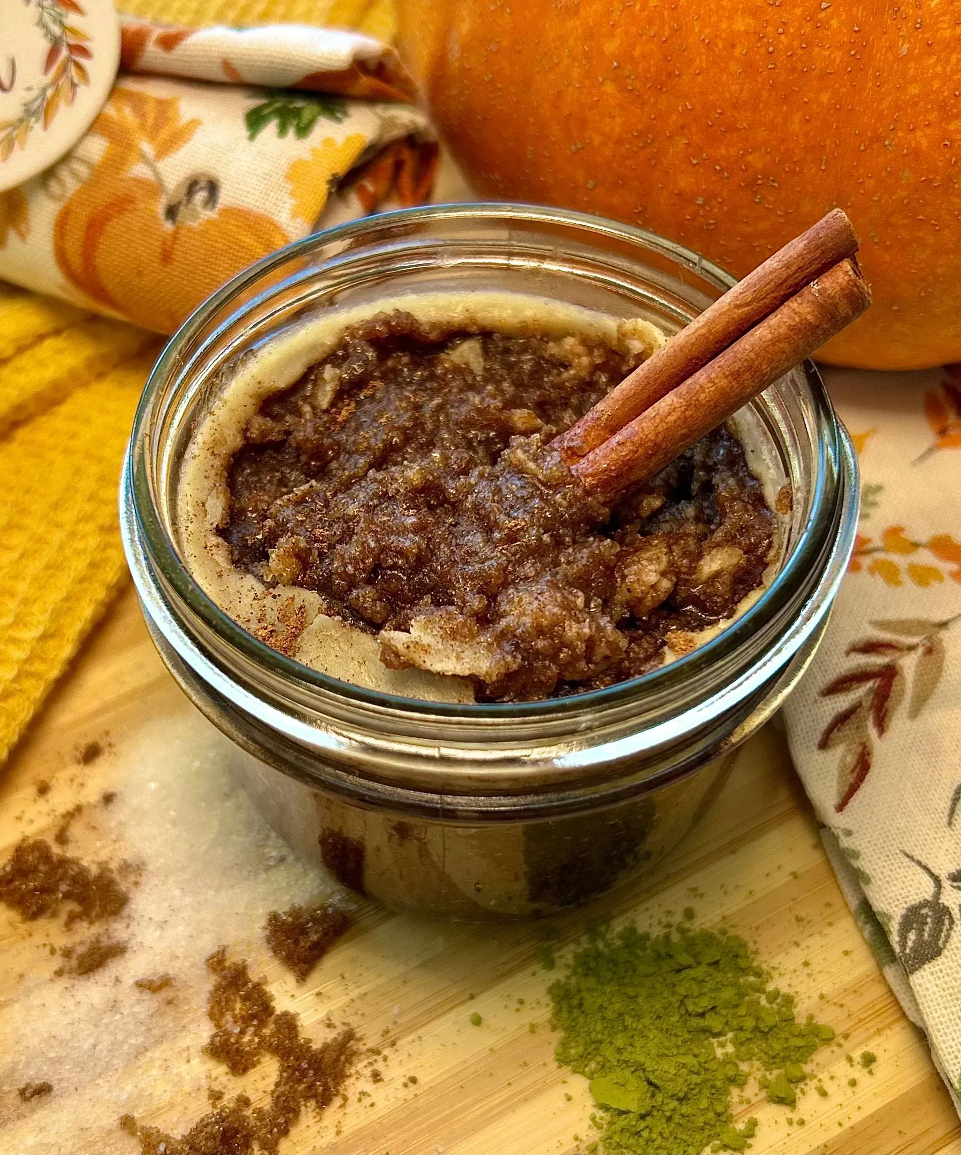 A glass jar of pumpkin spice scrub with a cinnamon stick, resting on a wooden board with scattered spice and matcha powder.