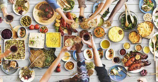People raising glasses in a toast around a table filled with diverse dishes of food, including olives, salads, and roasted vegetables.