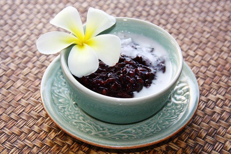 A bowl of black sticky rice with coconut milk, garnished with a white flower, on a green saucer.