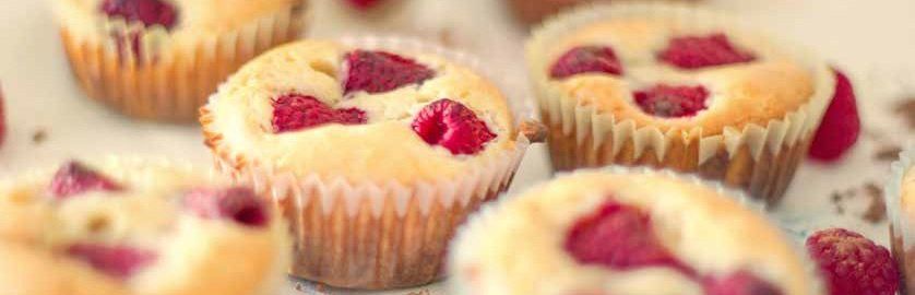 Close-up of several muffins with raspberries baked into the tops, in a white paper cup. The baked goods have golden brown tops.