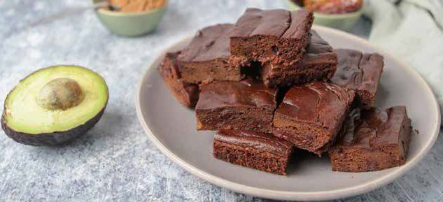 Brownie squares piled on a plate next to a sliced avocado. Bowls of brown powder sit in the background.