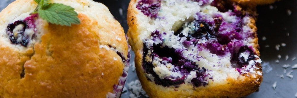 Close-up of two blueberry muffins, golden brown with visible blueberries and a garnish of mint.