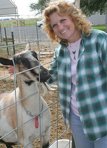 Woman in green plaid shirt petting a goat behind a wire fence, smiling outdoors.