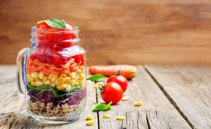 Rainbow salad in a jar with layered vegetables, sitting on a wooden table. Tomatoes, corn, carrots, and quinoa are visible.