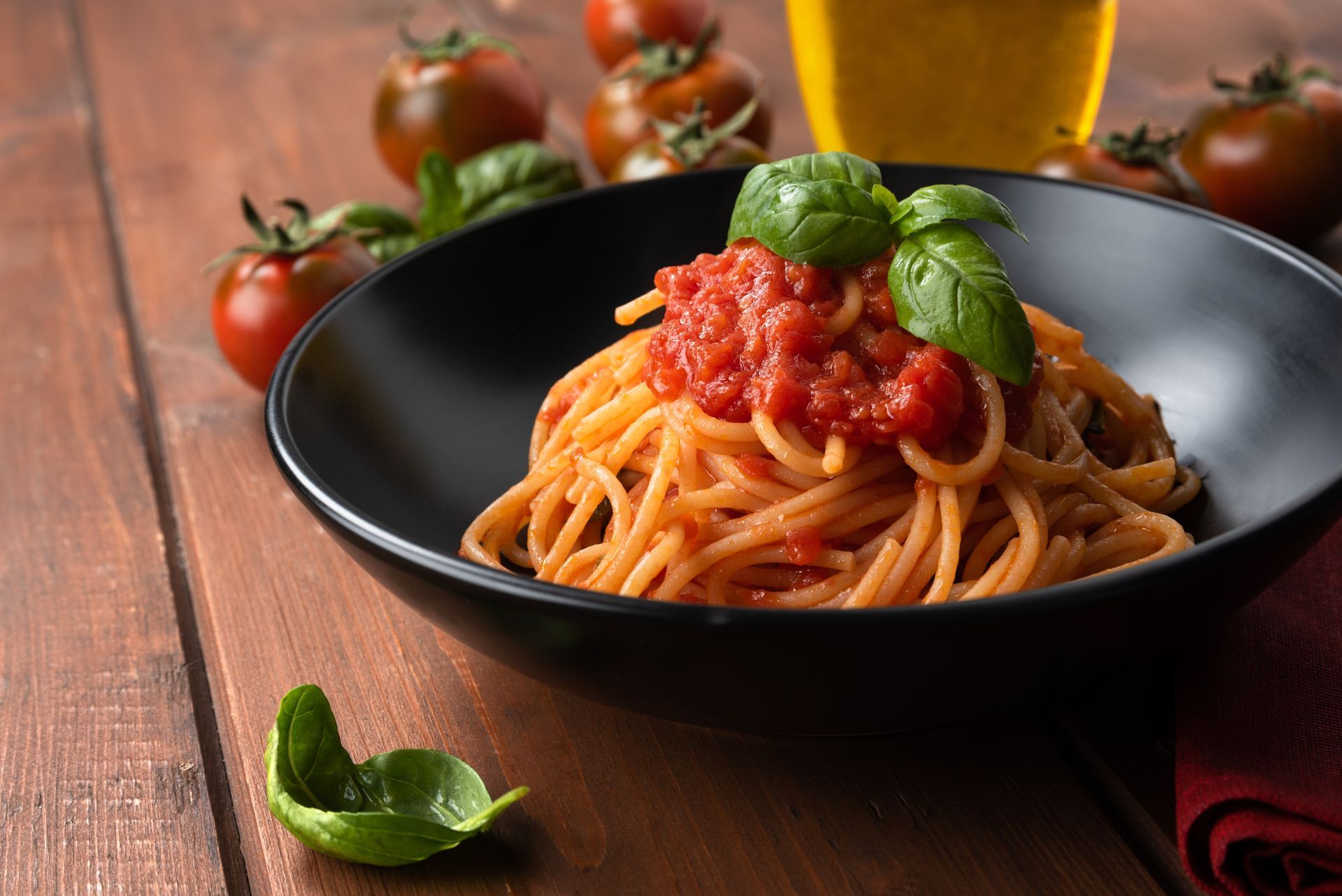 A black bowl of spaghetti topped with red tomato sauce and fresh basil leaves, set on a wooden table with cherry tomatoes.