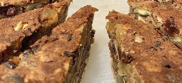 Close-up of sliced, brown loaf cake studded with raisins, nuts, and seeds. The cake is sitting on a white surface.