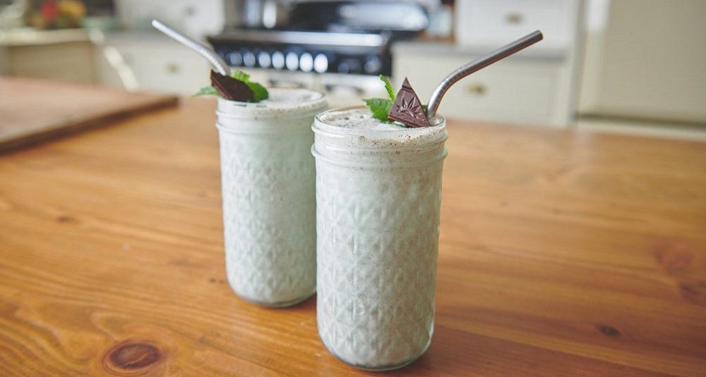 Two tall, light green smoothies in glass jars with metal straws, garnished with mint and chocolate, on a wooden table.
