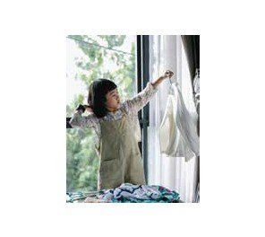 A young girl wearing overalls is hanging laundry by a window. She holds up a white garment, with a pile of clothes on the table in front of her.