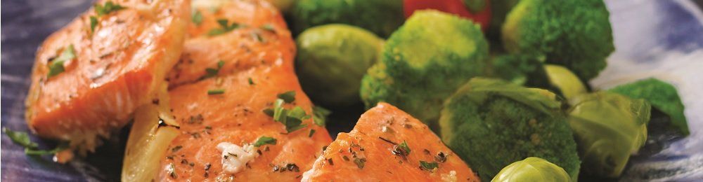 Salmon fillets with green vegetables on a plate. The salmon is a vibrant orange with herbs. The green vegetables are likely Brussels sprouts.