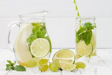 A pitcher and glass of lime and mint infused water, with grapes and ice on a white wooden surface.
