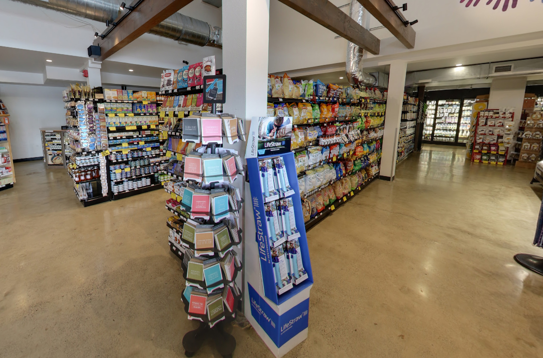 A brightly lit convenience store interior showing aisles of packaged snacks, a greeting card display, and tile flooring.