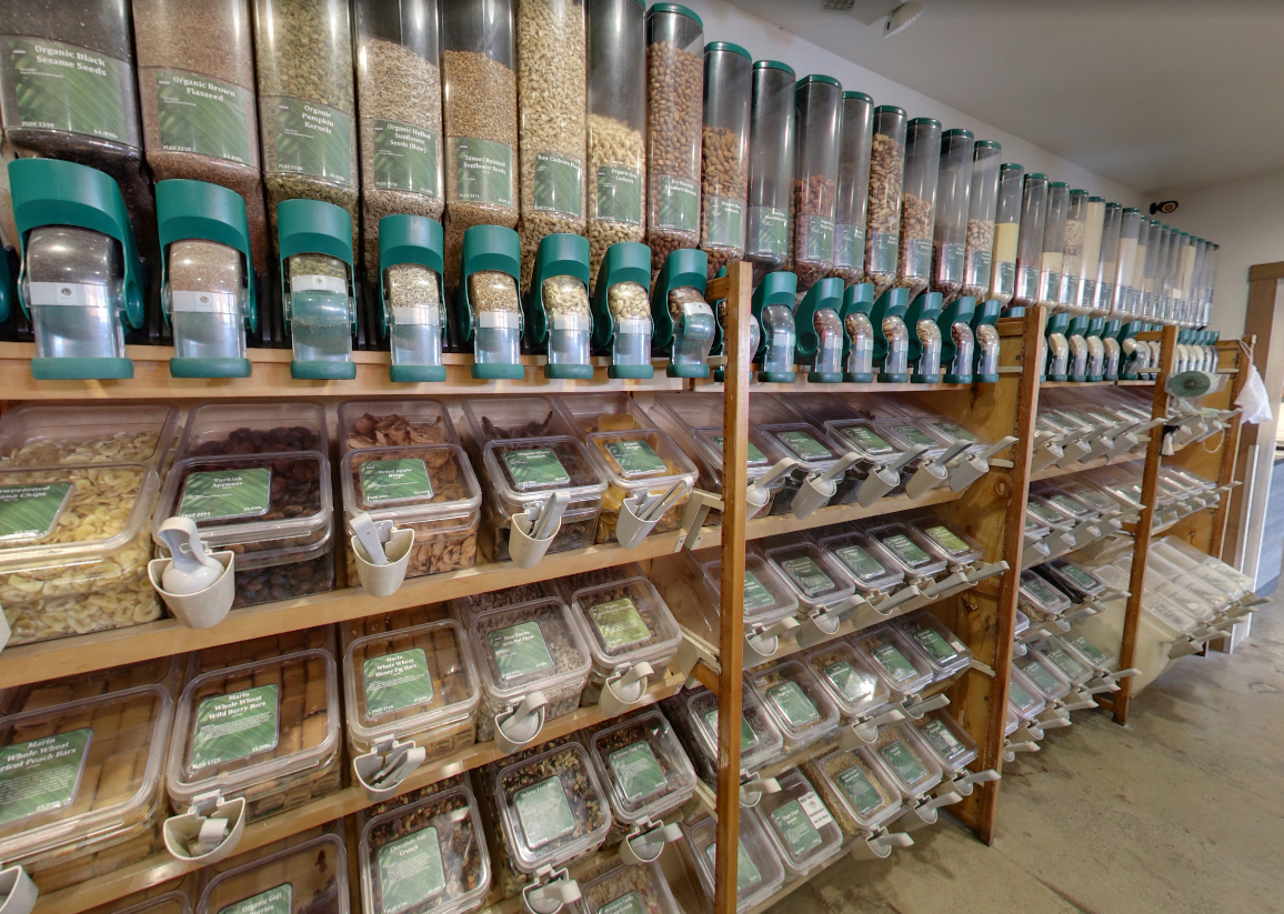 Bulk food store shelves stocked with clear containers of dry goods and gravity dispensers with green handles.