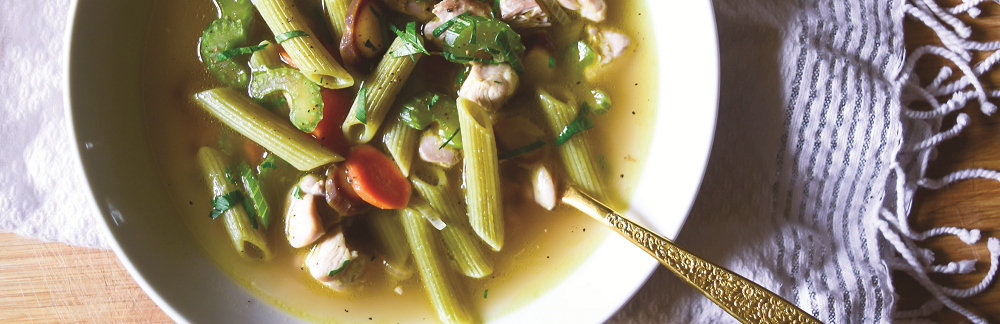 A bowl of soup with penne pasta, chicken, and vegetables, served with a golden spoon, on a wooden table with a patterned napkin.