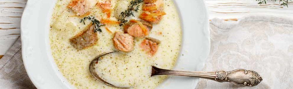 Creamy soup with salmon chunks, herbs, and a silver spoon in a white bowl on a table.