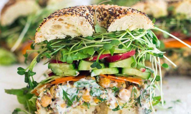 Everything bagel sandwich overflowing with avocado, sprouts, and colorful vegetables.