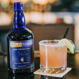 A blue Wild Tonic Jun Kombucha bottle next to a cocktail in a glass rimmed with salt and a lime wedge.