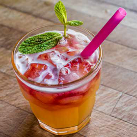 A colorful cocktail in a glass with ice, a pink straw, and mint leaves. The drink is orange with red and white layers.
