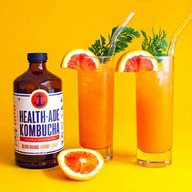 A bottle of Health-Ade kombucha next to two tall glasses of orange kombucha drinks, garnished with orange slices and green sprigs.