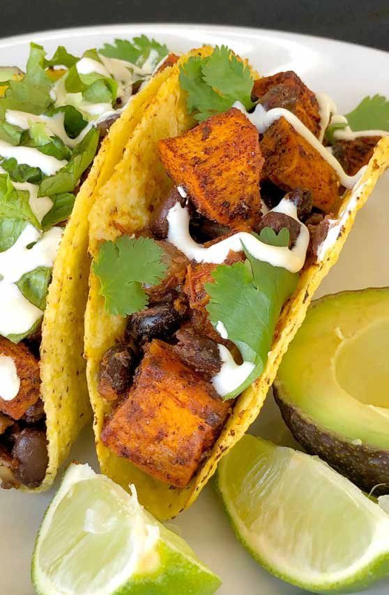 Close-up of two hard-shell tacos filled with sweet potatoes, black beans, cilantro, and white sauce, served with avocado and lime.