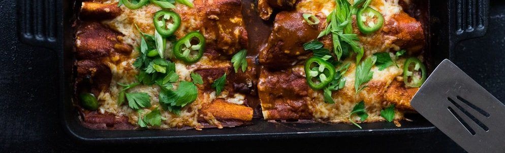 Overhead shot of a casserole with sliced jalapenos and cilantro garnish, served in a dark rectangular dish. A spatula rests at the right edge.