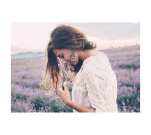 Woman in white dress smelling lavender in a field. Purple flowers, earrings, and sunlight visible.