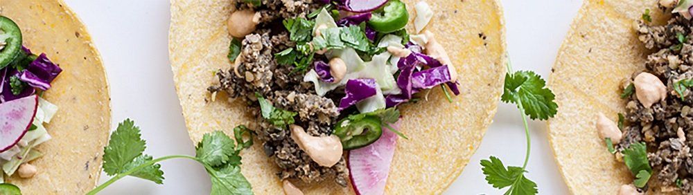 Three tacos on a white surface; each filled with different toppings, including meat or mushrooms, greens, red cabbage, and red onion.