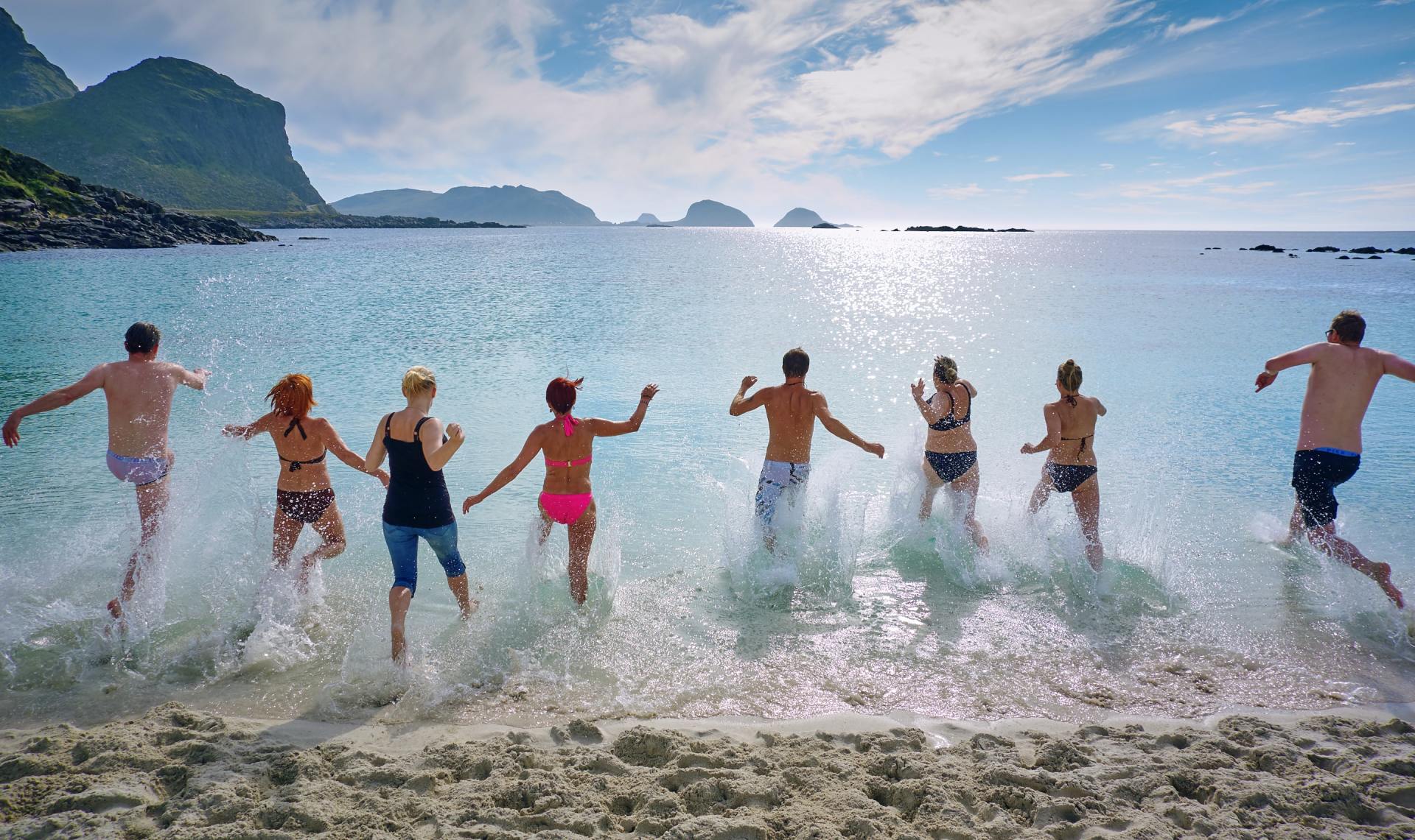 Group of people jumping into the ocean on a sunny day; the water is splashing as they run.