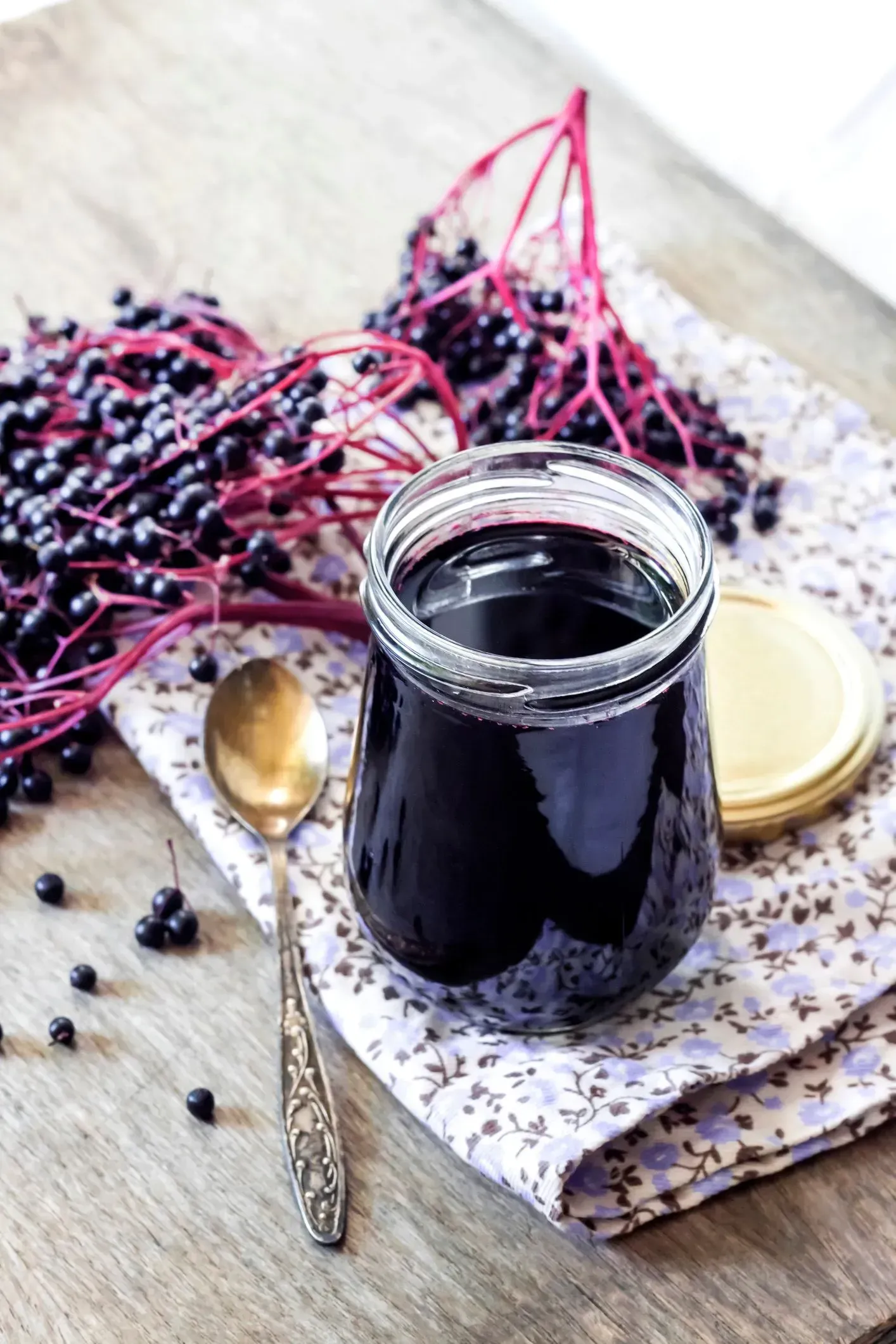 A glass jar of dark elderberry syrup, a silver spoon, and clusters of fresh elderberries on a floral napkin and wood table.