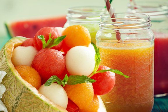 A cantaloupe bowl filled with melon balls, next to fruit juice in a jar with a straw, and watermelon slices.