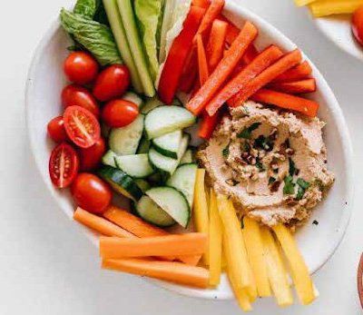 A white platter with colorful vegetables, including carrots, cucumbers, tomatoes, and hummus.