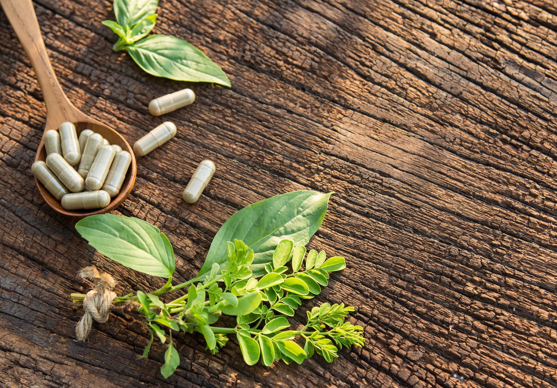 Wooden spoon with capsules and fresh herbs on a weathered wooden surface.