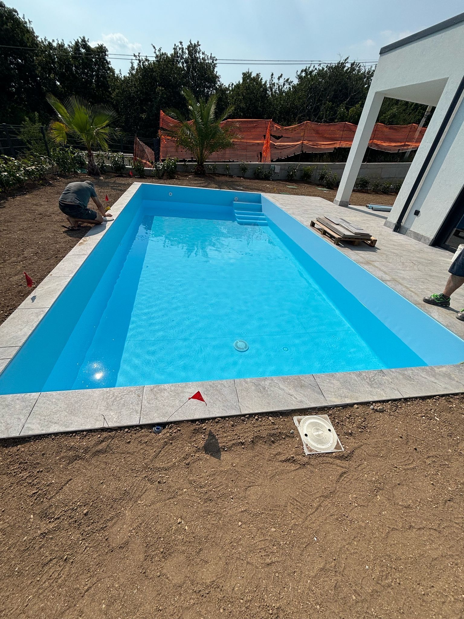 Piscina rettangolare blu accanto a una casa moderna, con ghiaia e alberi, immersa nella luce del sole.