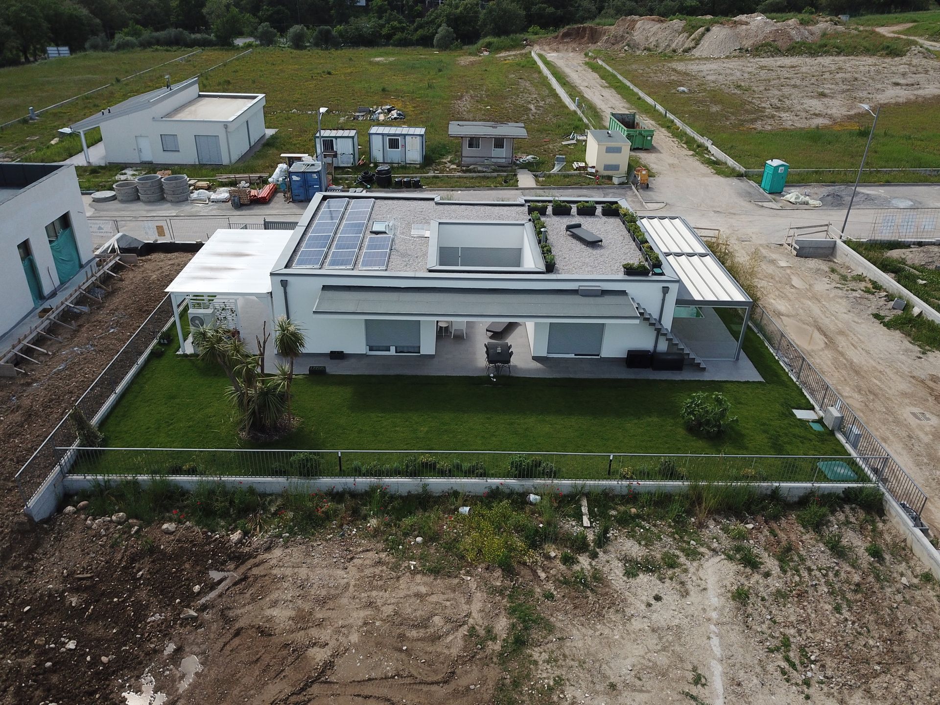 Veduta aerea di una casa bianca con tetto piano e prato verde, circondata da strade sterrate e attrezzature da cantiere.