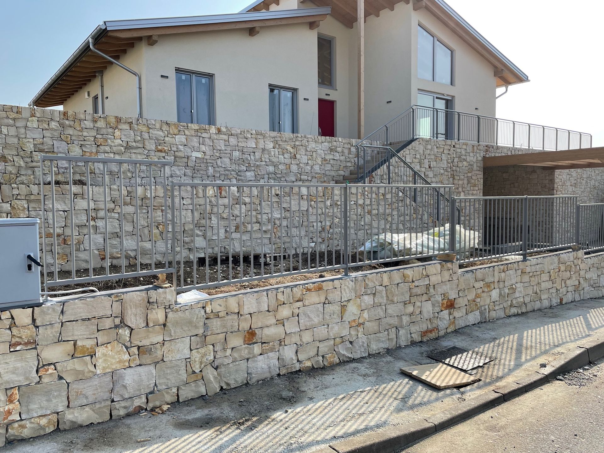 Casa in pietra dietro un basso muro di pietra e una recinzione metallica accanto a una strada asfaltata, attualmente in costruzione.