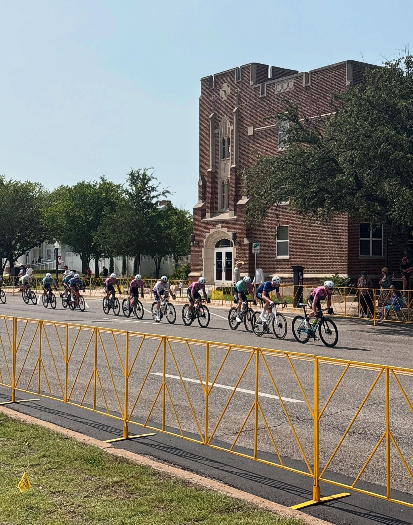 Cyclists in a race pass a brick building. Yellow barriers line the road.