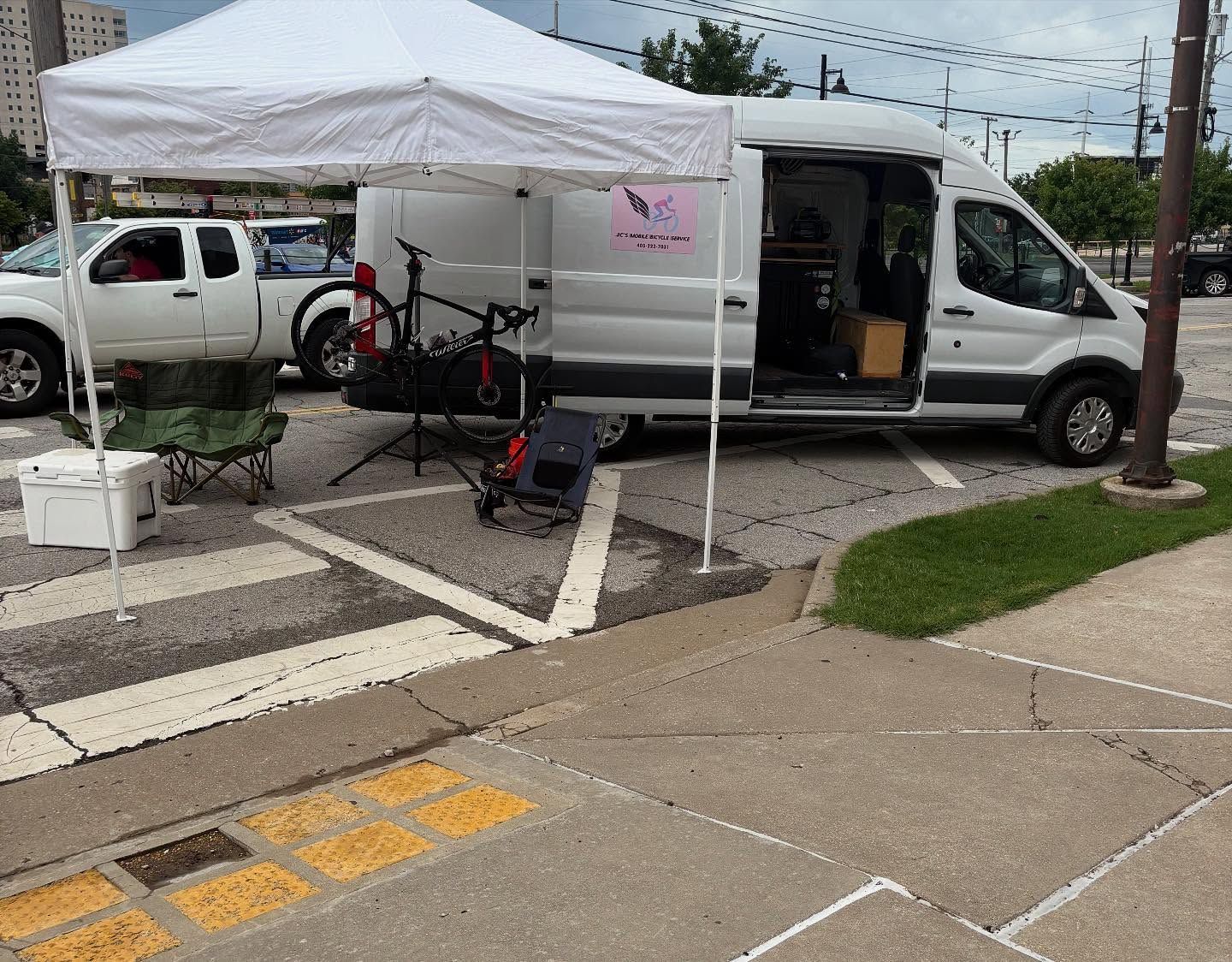 White van parked under a canopy in a parking lot. A white pickup truck is visible. A bike and chairs are nearby.