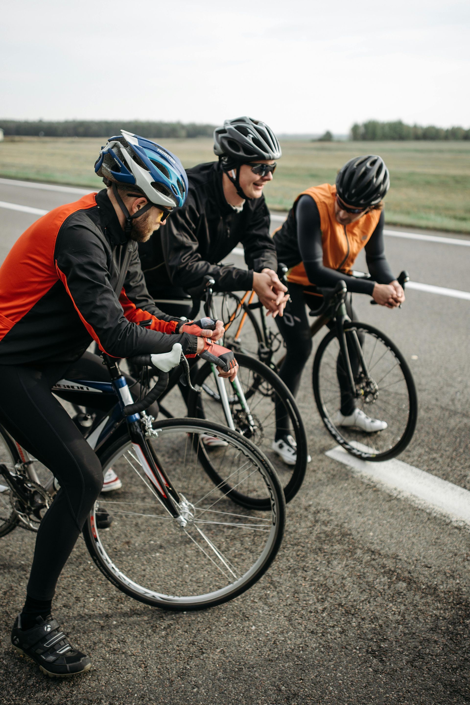 Three cyclists in helmets and gear, resting on road, leaning on bikes, smiling.