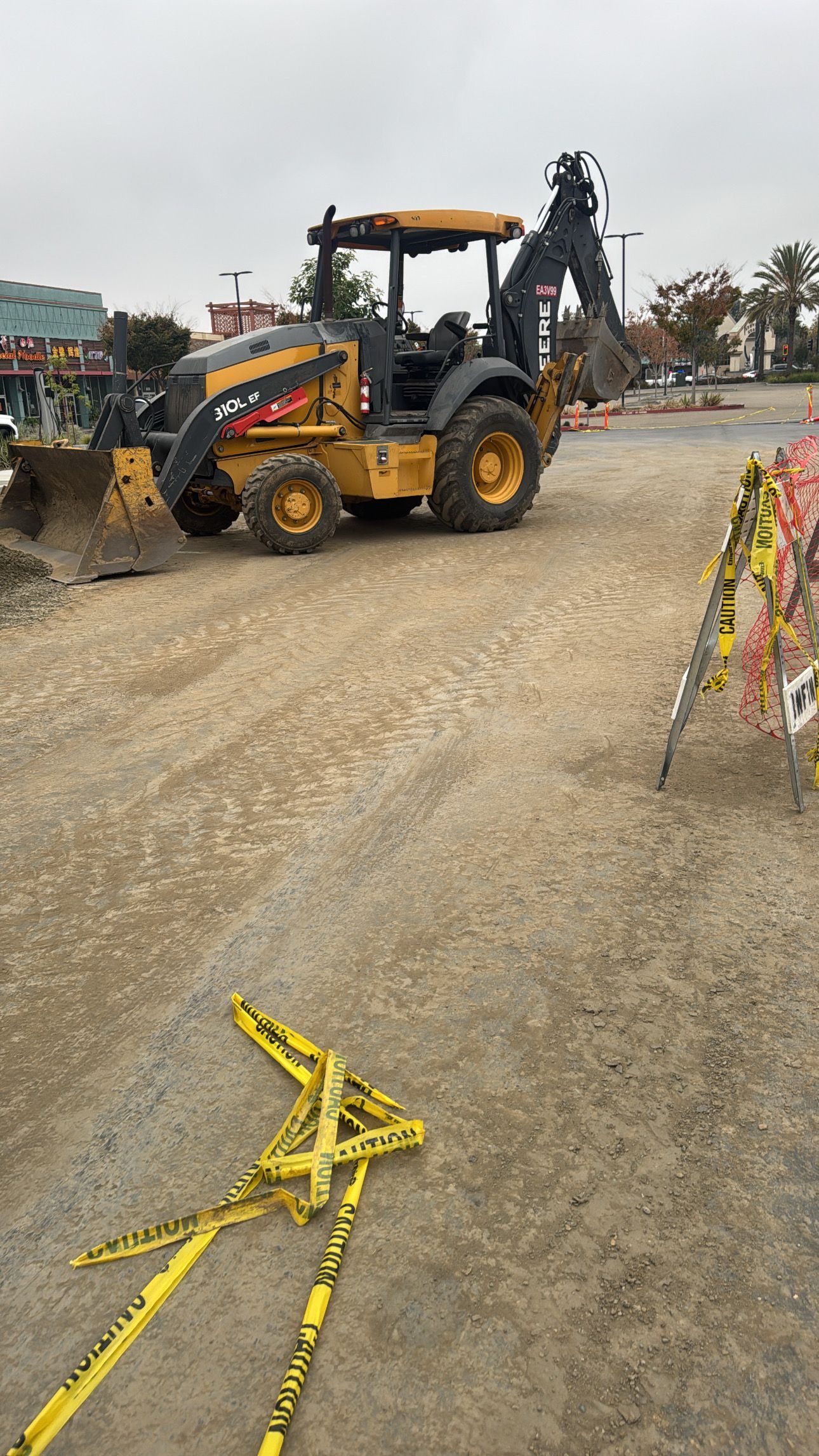 Yellow construction backhoe on a dirt lot, with caution tape in the foreground. Overcast sky.