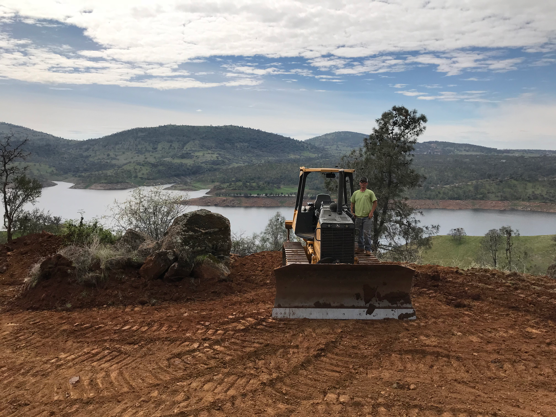 Dozer clearing land overlooking a lake and hills, operator visible.