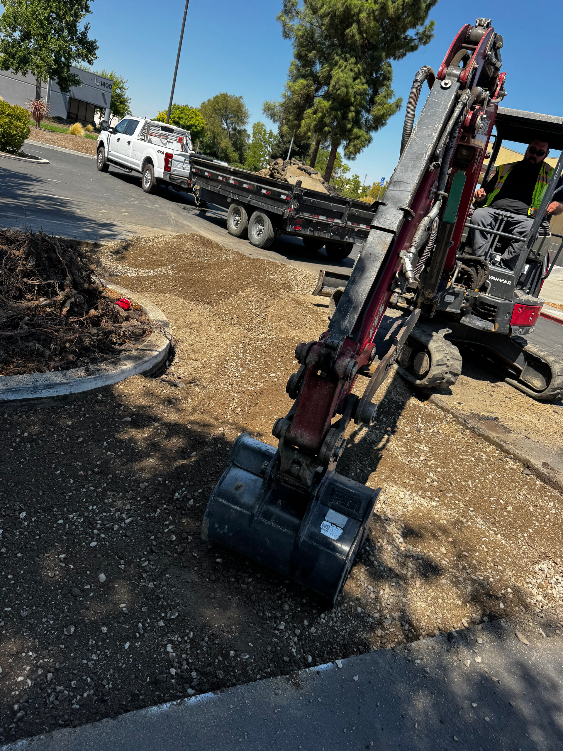 Excavator removing asphalt on a sunny day. A white truck and trailer are parked nearby.