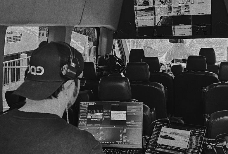 Inside a van, a person wearing a headset operates a laptop displaying video editing software, with multiple screens visible.