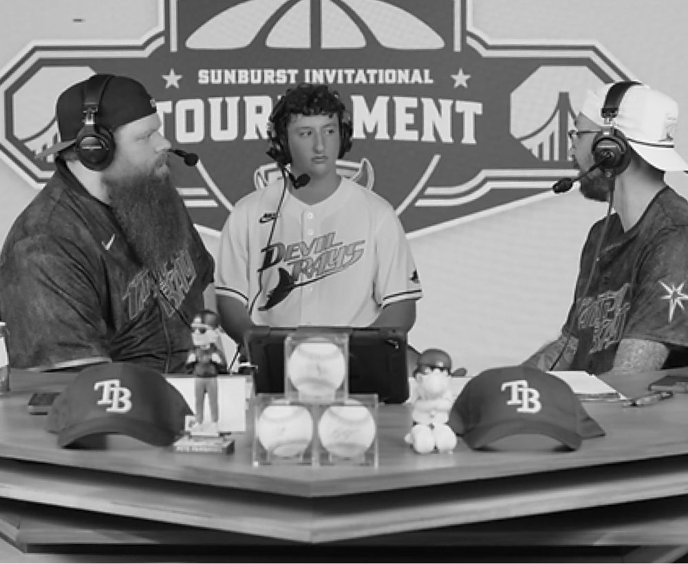 Three people at a table, two wearing baseball jerseys, with microphones. Baseball-themed props in front of them.