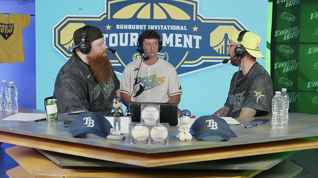 Three men at a baseball-themed tournament desk, discussing; two wear headsets. Tampa Bay Rays logos on caps and jerseys.